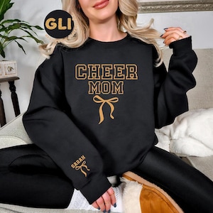 Könnte beinhalten: Schwarzes Sweatshirt mit dem Text "CHEER MOM" in goldfarbenen Buchstaben und einer goldenen Schleifengrafik. Der Name "SARAH" ist in Gold auf dem linken Ärmel aufgedruckt. Das Sweatshirt ist ein lässiger Stil.