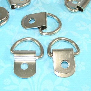 May include: Five silver metal D-rings with a flat back and a hole in the center. The D-rings are on a blue background.