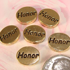 May include: Gold-colored, round charms with the word "Honor" in black lettering. The charms are slightly domed and have a metallic sheen. A U.S. dime is included for size comparison. The charms are likely for jewelry making or crafting.
