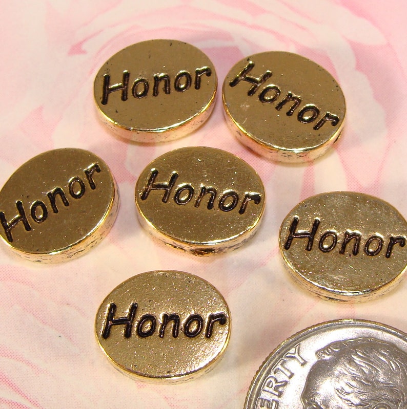 May include: Gold-colored, round charms with the word "Honor" in black lettering. The charms are slightly domed and have a metallic sheen. A U.S. dime is included for size comparison. The charms are likely for jewelry making or crafting.