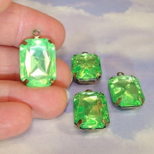 May include: Four green glass emerald-shaped pendants with silver settings. The pendants are on a white surface.