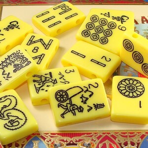 May include: A set of yellow mahjong tiles with black markings. The tiles feature various symbols and numbers, including a dragon, a wheelbarrow, and a flower. The tiles are arranged in a random pattern on a red and gold surface.