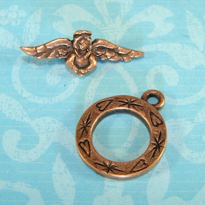 Op de afbeelding: Twee antieke bronzen bedels, een is een gevleugelde engel met een halo en de andere is een ronde bedel met een hart- en stervormig ontwerp.