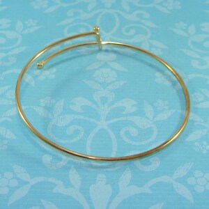 Puede incluir: Un brazalete de oro con un diseño simple y minimalista. El brazalete está abierto y tiene un cierre con bisagra.