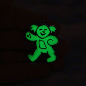 May include: A luminous green bear-shaped pendant with a black outline. The bear is depicted walking and smiling. The pendant has a small loop for hanging. The background is dark.