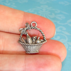 May include: Silver charm of a wicker basket filled with a bottle of wine, cheese, and fruit. The basket has a bow on top and a loop for hanging.