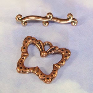 May include: Two copper-coloured metal charms. One charm is a simple, curved line with decorative swirls at each end. The other charm is shaped like a butterfly with a detailed, ornate design.