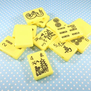 May include: A set of yellow mahjong tiles with black Chinese characters and symbols. The tiles are arranged in a pile, with some of the tiles overlapping. The tiles are made of a smooth, glossy material.