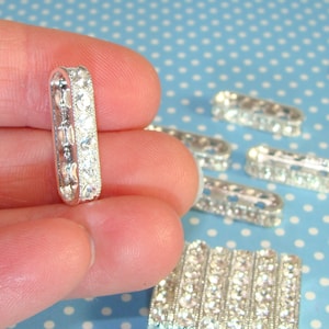 May include: Silver-toned metal slider with clear rhinestones. The slider is rectangular and has a rounded edge. The slider is shown in a hand, along with several other sliders.