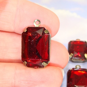 Puede incluir: Primer plano de tres colgantes de cristal rojo con engastes de metal dorado. Los colgantes son de forma rectangular y tienen un diseño facetado. Los colgantes se sostienen en una mano.