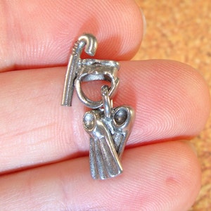 Puede incluir: Un pequeño dije plateado con un diseño estilizado. El dije está compuesto por múltiples elementos, incluyendo una parte superior curvada, un anillo central y una sección inferior con apariencia drapeada. El dije se sostiene entre los dedos.