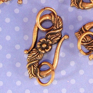 Puede incluir: Primer plano de varios cierres decorativos de color bronce. Cada cierre presenta un diseño floral con una flor central y detalles de hojas. Los cierres tienen un cierre de gancho y ojo y están colocados sobre un fondo azul con lunares blancos.