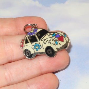 May include: A cream-colored car charm with black windows and wheels. The car is decorated with blue, purple, and red floral and heart designs. The charm has a silver-colored metal border and a small loop for attaching to a keychain or necklace.