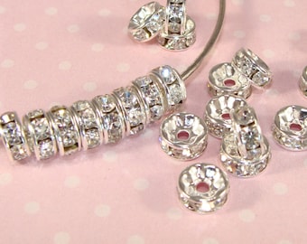 May include: Silver-toned metal bead caps with clear rhinestones. The bead caps are arranged in a cluster on a pink background.