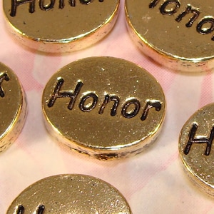 May include: Close-up of several gold-colored, round charms. Each charm has the word "Honor" in black, embossed lettering. The charms are scattered on a light pink and white patterned surface.