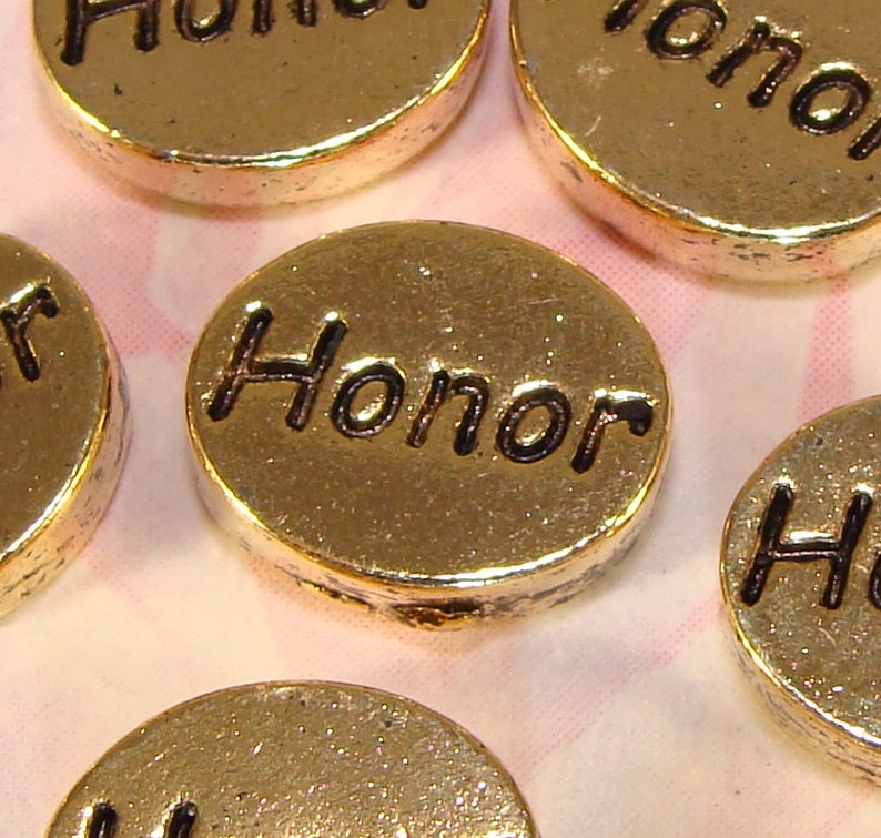 May include: Close-up of several gold-colored, round charms. Each charm has the word "Honor" in black, embossed lettering. The charms are scattered on a light pink and white patterned surface.