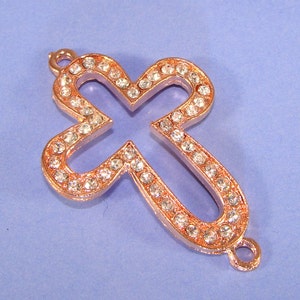 Op de afbeelding: Een roosgoudkleurige kruisbedel met heldere strasssteentjes. Het kruis is in het midden open en heeft bovenaan een lus om aan een ketting of armband te bevestigen.