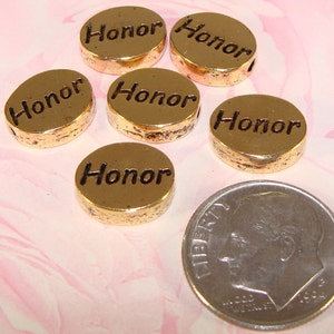 May include: Six gold-colored, round beads with the word "Honor" engraved on them. The beads are next to a dime for size comparison. The beads have a slightly distressed finish and a hole for stringing.