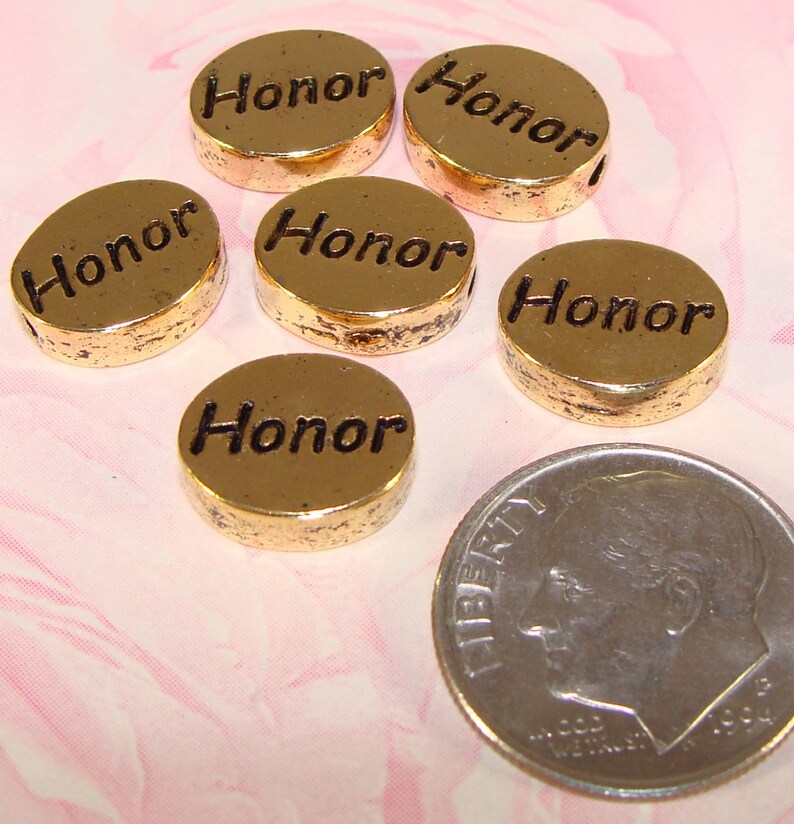 May include: Six gold-colored, round beads with the word "Honor" engraved on them. The beads are next to a dime for size comparison. The beads have a slightly distressed finish and a hole for stringing.