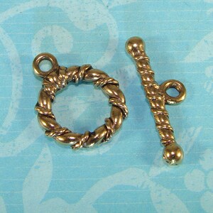 Può includere: Un fermaglio a bottone in metallo dorato con un design a corda intrecciata. Il fermaglio ha un anello rotondo a un'estremità e una barra dritta con un'estremità rotonda all'altra.