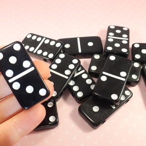 Può includere: Una collezione di domino neri con punti bianchi. I domino sono disposti in una pila, con alcuni di essi sovrapposti. I domino sono fatti di un materiale nero liscio.