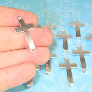 May include: Silver-toned metal cross charms with a loop at the top and bottom. The charms are arranged on a light blue background.