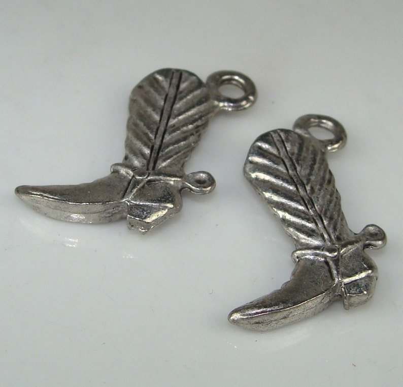 May include: Two silver-toned metal boot charms with a feather design. The boots have a pointed toe and a spur on the heel.