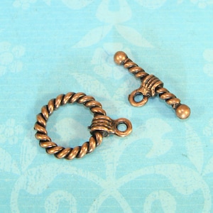 Puede incluir: Dos cierres de palanca de tono cobre antiguo con un diseño de cuerda. El cierre de la izquierda es un círculo con un pequeño bucle en el lateral. El cierre de la derecha es una barra con un bucle en el medio.