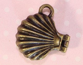 May include: A bronze-colored seashell charm with a hinged design and a loop for attaching to jewelry or other crafts. The shell has a detailed, ridged texture and a slightly aged appearance, suggesting a vintage or antique style. The charm is shown against a pink background.