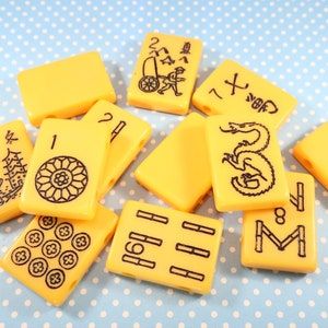 May include: A set of yellow plastic mahjong tiles with black markings. The tiles feature various symbols and numbers, including a dragon, bamboo, and circles.