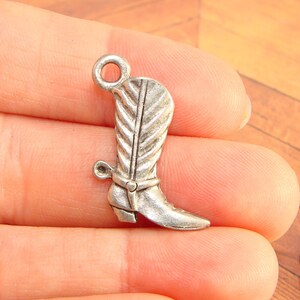 May include: Silver charm in the shape of a cowboy boot with a spur. The boot has a decorative stitching pattern and a loop at the top for attaching to a necklace or bracelet.