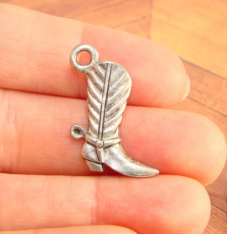 May include: Silver charm in the shape of a cowboy boot with a spur. The boot has a decorative stitching pattern and a loop at the top for attaching to a necklace or bracelet.