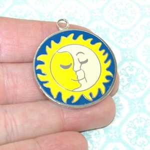 Può includere: Un ciondolo rotondo con bordo color argento. Raffigura un sole giallo e una luna color crema, entrambi con gli occhi chiusi, su sfondo blu. Il sole e la luna si toccano. Il ciondolo ha un piccolo anello nella parte superiore.