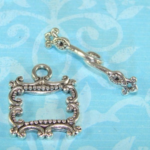 May include: Two silver-toned metal embellishments. One is a small ornate frame with a square opening. The other is a decorative, curved piece with a small, round, raised detail.