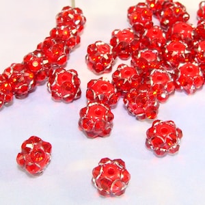 May include: A close-up of a group of red rhinestone beads. The beads are round and have a faceted design. They are scattered on a white surface.