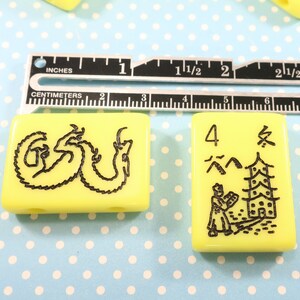 May include: Two yellow plastic mahjong tiles with black ink. The first tile has a black dragon design. The second tile has a black illustration of a person standing in front of a pagoda with the number 4 and the Chinese character for "wind" written in black ink.