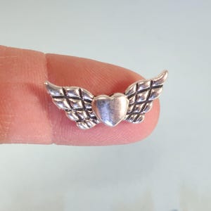 May include: A small, silver-colored charm featuring a heart with textured wings. The wings have a detailed, cross-hatched pattern. The charm is held between two fingers, with a light blue background.