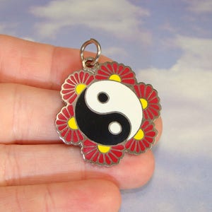May include: A silver pendant with a yin-yang symbol in black and white at its core, encircled by red and yellow floral designs. It has a small loop for attachment. The background is a light blue sky.
