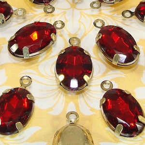 May include: A group of ten oval-shaped red glass rhinestones with silver settings and gold jump rings. The rhinestones are arranged in a circular pattern on a white and gold patterned fabric.
