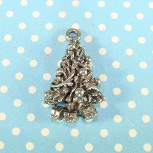 Puede incluir: Un amuleto de plata con forma de &aacute;rbol de Navidad con adornos y regalos. El amuleto est&aacute; sobre un fondo azul con lunares blancos.
