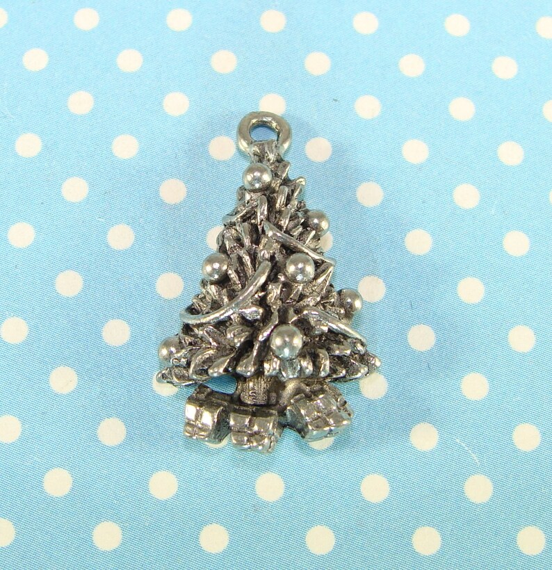 Puede incluir: Un amuleto de plata con forma de &aacute;rbol de Navidad con adornos y regalos. El amuleto est&aacute; sobre un fondo azul con lunares blancos.