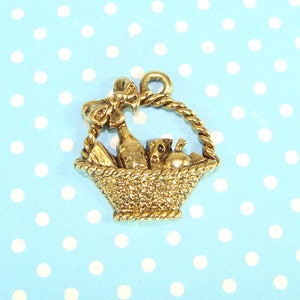May include: Gold-toned metal charm shaped like a basket filled with a bottle of wine, a loaf of bread, a block of cheese, and an apple. The basket has a bow on top and a loop for hanging.