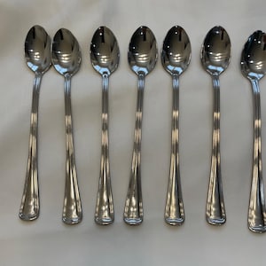 Op de afbeelding: Zeven roestvrijstalen lepels met lange handvatten en ovale kommen zijn gerangschikt op een wit oppervlak. De lepels hebben een reflecterende, gepolijste afwerking en de handvatten hebben een subtiel, geribbeld ontwerp. De lepels zijn waarschijnlijk om te serveren of te roeren.