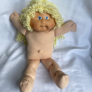 May include: A vintage Cabbage Patch Kid doll with a soft, flesh-toned body and limbs. The doll has a round face with blue eyes, rosy cheeks, and a small mouth. Its light yellow yarn hair is styled in a curly fashion. The doll is lying on a white surface.