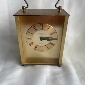 Puede incluir: Un reloj de cuarzo Seth Thomas dorado con asa. La esfera del reloj tiene números romanos y manecillas negras. El reloj es rectangular con líneas verticales en los lados y una pequeña base.