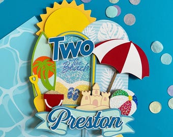 May include: A beach-themed cake topper with a yellow sun, surfboard, and a red and white striped umbrella. The topper includes the words "Two" and "Preston", along with a sandcastle, beach ball, and sand pail.