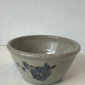 May include: A speckled ceramic bowl in a light colour with a flared shape. The bowl has a hand-painted design of blue flowers and brown stems. The interior of the bowl is a solid, light colour.
