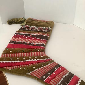 May include: A Christmas stocking featuring horizontal bands of olive green velvet, red satin, and brown fabric. The stocking is decorated with silver jingle bells, silver rickrack trim, small embroidered stars, and scattered sequins in red, green, and silver.
