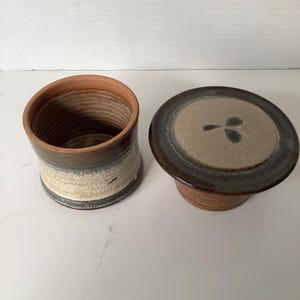 May include: A ceramic butter crock consisting of a cylindrical base with a terracotta interior and a two-toned brown and cream exterior glaze, paired with a matching lid featuring a flat, speckled beige top with a dark three-leaf painted design.