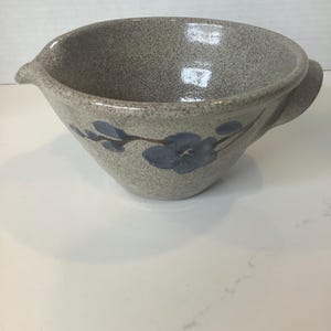 May include: A speckled gray ceramic mixing bowl with a pouring spout and handle. The bowl features a blue floral design. The bowl is sitting on a white surface.